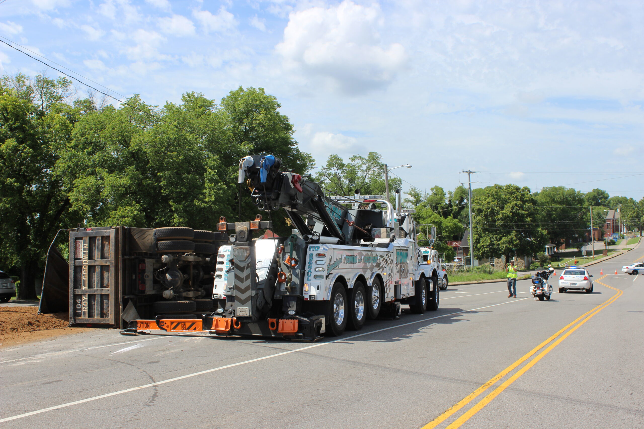 A tow pro truck on the side of the road with the road closed down by police officers. Off the road is an over turned garbage truck that needs to be recovered.