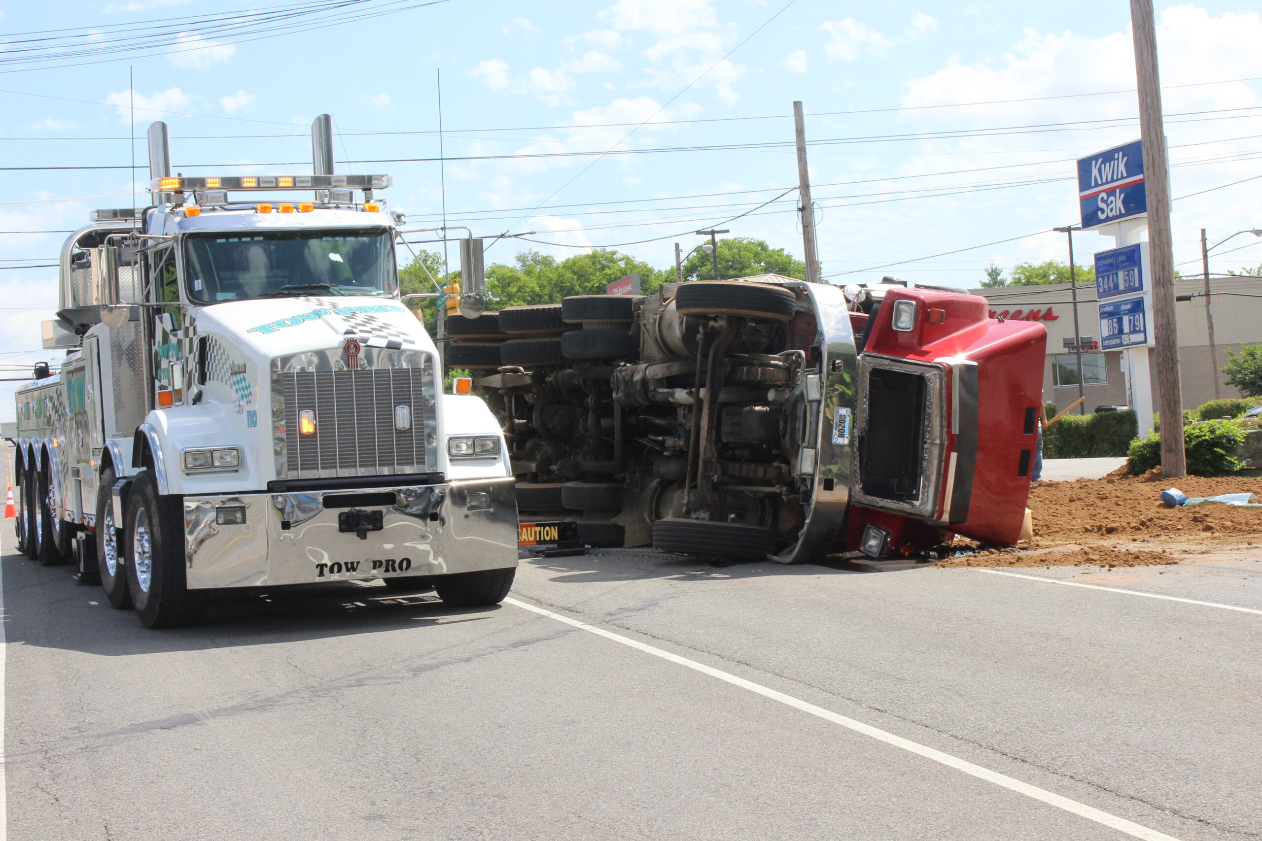 A tow pro truck pulled to the side of the road next to a over turned red semi-truck.